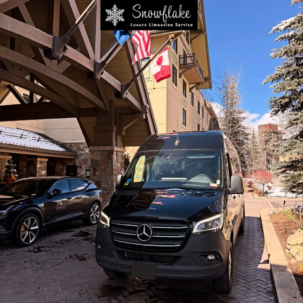 A black Mercedes-Benz Sprinter luxury shuttle van is parked at the covered entrance of a mountain resort or hotel on a sunny winter day. Snow-covered trees line the walkway, and U.S. and Canadian flags hang above the entrance. A sign reading “Snowflake Luxury Limousine Service” appears at the top of the image, while another dark SUV is parked nearby. The scene suggests premium transportation service in a ski resort setting.
