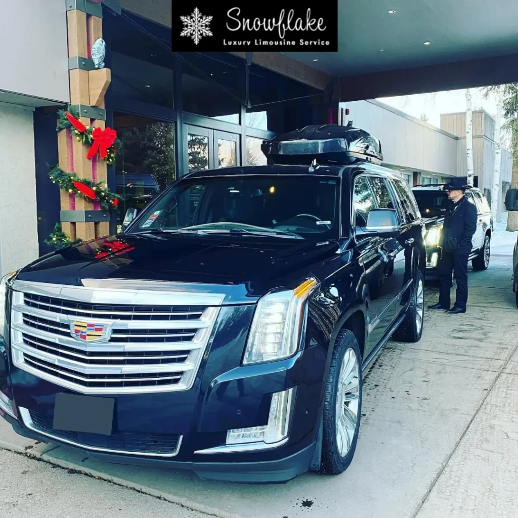 **Alt text:** A black Cadillac Escalade SUV is parked at the covered entrance of an upscale hotel or resort decorated with holiday wreaths and red bows. A uniformed chauffeur stands beside a second luxury SUV waiting behind it. The lead vehicle is equipped with a rooftop cargo carrier, suggesting airport or resort transfers with luggage. A “Snowflake Luxury Limousine Service” logo banner appears at the top of the image, emphasizing premium chauffeured transportation.